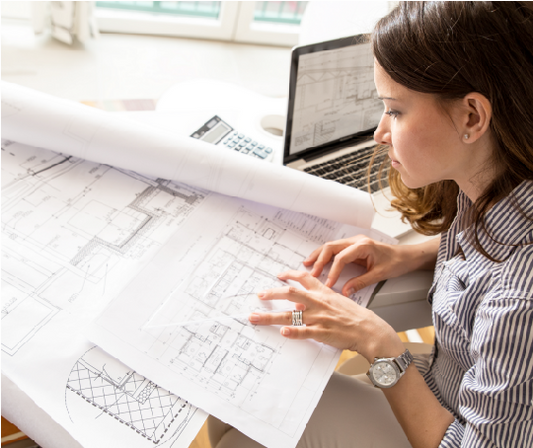 Woman working on architectural blueprints with a laptop at a desk.